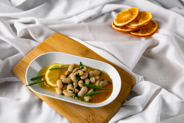 Marinated white beans in their own juice on an oval plate with Thai pineapple sauce and green onions. Decorated with a slice of lemon and dried oranges on a wooden Board with a snow white fabric