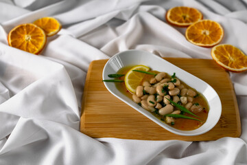 Marinated white beans in their own juice on an oval plate with Thai pineapple sauce and green onions. Decorated with a slice of lemon and dried oranges on a wooden Board with a snow white fabric