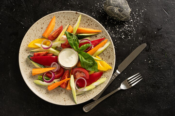 Vegetable salad of yellow and red pepper, carrots, fresh cucumber, onions and basil with sauce. A plate of fresh veggie salad on a black table. Top view.