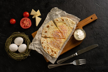 A wheat tortilla stuffed with cheese and tomatoes cut on a cutting wooden board next to sour cream, cutlery, and ingredients on a black table. Top view.