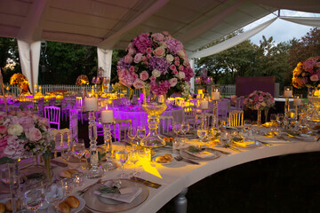 A luxury alfresco wedding reception set up at the countryside; white rustic tiffany chairs, top table flower arrangements, crystal center pieces and candles.
