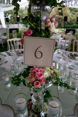 Table number stand of a luxury alfresco wedding reception at the countryside; white rustic tiffany chairs, top table flower arrangements and crystal center pieces.