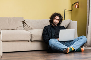 Low angle view of handsome curly man looking at camera while using laptop on floor in living room