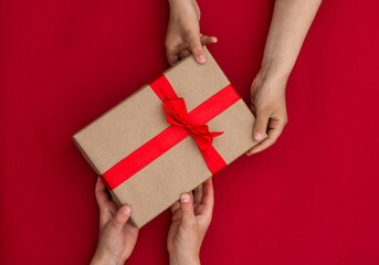Hands giving or receiving a gift in craft paper with a red ribbon on red background. Top view. Copy...
