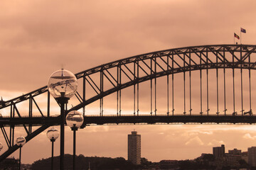 Sydney harbour bridge with the Opera House's lamp posts in the foreground. Blues point tower can be seen in the distance. Orange cloudy sky, dusk, sunset. Cityscape