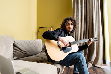 Selective focus of handsome man playing acoustic guitar near laptop on couch