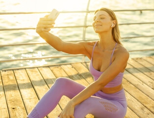 Morning workout. Young woman in sportwear with earphones doing selfie portrait on smartphone on the beach at sunrise