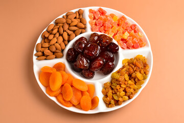 Mix of dried fruits and nuts on a white plate. Apricot, almond, raisin, dates fruit. On a brown background. Space for text or design.