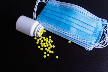 Scattered yellowish round tablets, vitamins scattered from a white plastic bottle, and a blue protective medical face mask on a black background. Prevention of covind-19 or coronavirus. Close up