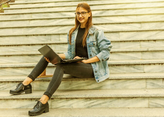 Fototapeta na wymiar Happy modern woman student in denim jacket sitting on the stairs with laptop. Distance learning. Modern youth concept.