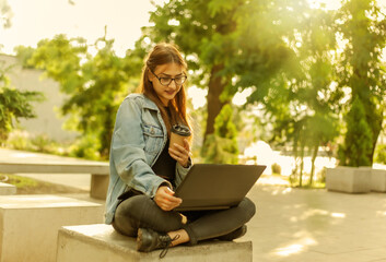 Young modern woman student in a denim jacket sitting at park and looks at the laptop screen with coffee cup on hand. Distance learning. Modern youth concept.