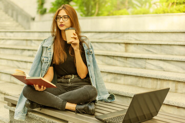 Modern students. Distance learning. Young hipster girl reads book while sitting on bench with a laptop and drink coffee