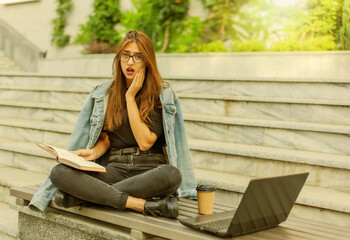 Modern students. Distance learning. Young enthusiastic woman reads book while sitting on bench with a laptop