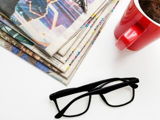 A stack of old Newspapers, black eyeglasses, a pen, and a red Cup of black coffee on a white...