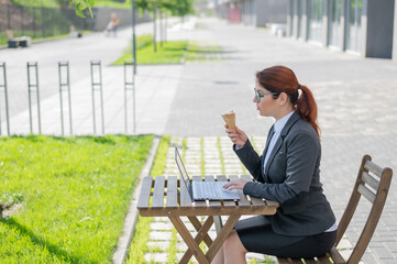 Fototapeta premium Happy woman typing on the computer and eating ice cream cone while sitting at a wooden table outdoors. Female employee remotely working on a laptop. Lunch break in a street cafe.