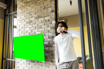 young man in pajamas drinking coffee while standing near green lcd screen hanging on brick wall
