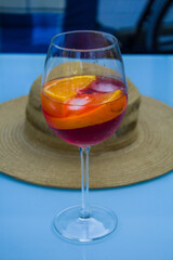 Red drink on light blue background, refreshing martini in glass cup with oranges and ice on a table outdoors with a straw hat in the background.