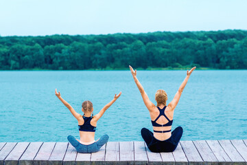 Mother and daughter doing yoga at the shore of the lake in summer.