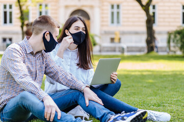 Student couple in facial masks studying outside on the grass, sitting down on the school campus...