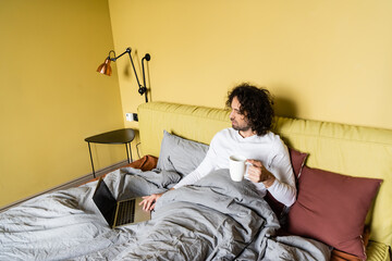high angle view of curly freelancer using laptop with blank screen while holding cup of coffee in bed