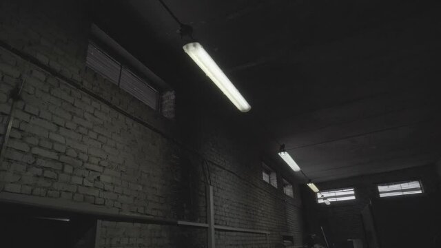 Fluorescent lights lamps turn on and turn off in dark room of plant, hangar, workshop department. Network electrical short circuit in industrial manufacturing area with brick walls and windows