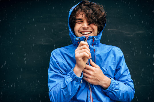 Portrait of the man smiling broadly, wearing blue raincoat during rain outside. Handsome male in blue raincoat enjoying the rain on black wall. Young man has joyful expression in rainy weather.