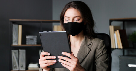 Businesswoman sitting at desk in office and using tablet device online. Beautiful female in mask tapping and scrolling on computer screen while browsing. Coronavirus pandemic.