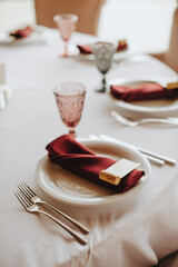 The table is covered with a white tablecloth, served. Plates, cutlery, napkin, pink glasses, seating cards, sprig of greens.