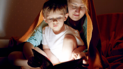 Portrait of cute little boy with young mother covering in blanket and reading a book with flashlight