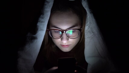 Fototapeta na wymiar A young woman lies under a blanket with a phone in her hands. A woman in the dark uses a pad. Modern electronic tablet. Close-up