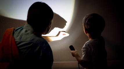 Closeup image of young mother teaching her little son showing shadows from torch on wall at night - obrazy, fototapety, plakaty