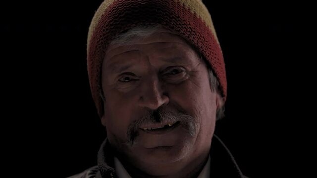 Close up. Hipster mature man in a rastaman hat nod his head and wink on camera in a semi lit light room isolated on a black background. Prores 422.