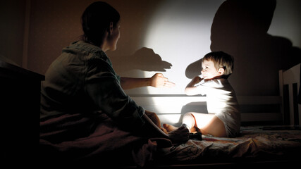 Happy smiling little boy and young mother playing with torch and shadows from their hands on wall at bedroom