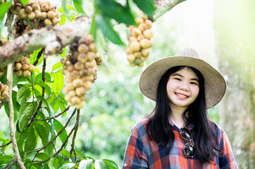 Happy young gardener lady wear plaid shirt and hat at his green organic garden enjoying the sunny day and longkong tree background.Sufficiency economy.Proud with success woman with organic orchards.