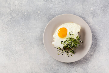 Traditional English breakfast with fried eggs with arugula microgreen in gray ceramic plate on gray concrete old background. Top view.