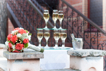 
A bride’s bouquet of orange roses next to champagne glasses set in tiers in daylight is shot in general.