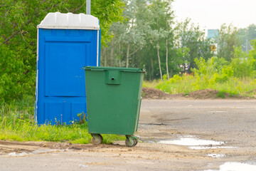Green trash can and portable toilet on a background of greenery