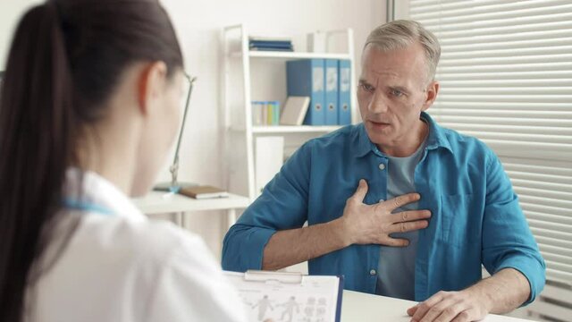 Rear view of middle-aged Caucasian man sitting at desk and describing symptoms of his illness to female physician wearing medical overall filling in patient card