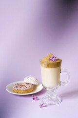 Beautiful dalgona drink a frothy coffee in a transparent mug and flowers lilac to purple Il purple background. Next to the mug is a marshmallow