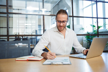 Photo of handsome young man making notes and working with laptop