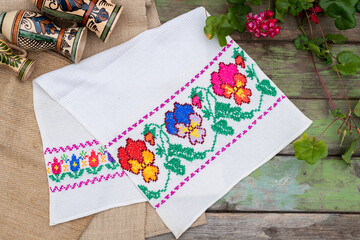 Handmade towel with colorful embriodered pattern with wooden background, canvas and folk art jugs and geranium flowers