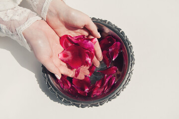 Dark pink peony petals in female hands and in bowl with clear water on white background. Organic cosmetics, spa concept. Copy space, flat lay, top view.