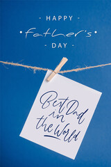 White greeting card with best dad in the world illustration hanging on rope with clothespins isolated on blue