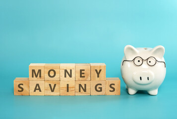 White ceramic piggy bank wearing reading glasses with wooden blocks spelling “MONEY SAVINGS” on blue teal background. Concept for money savings plan for retirement, aged society, financial accounting