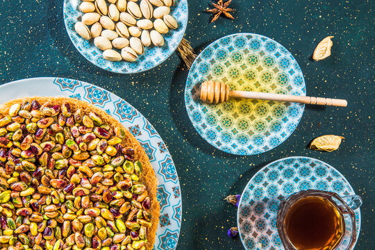 Top view of table with oriental dessert with pistachios and honey. Arabic sweet dessert of kadayif and pistachios on table with honey, wooden spoon and cup of tea.