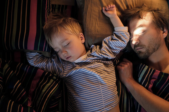 Cute toddler son and his father sleeping together on bed in bedroom at home. Real lifestyle family photo taken early morning. Fathers day concept.