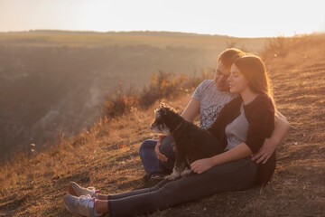 A young couple in love is sitting on a mountainside in the rays of the setting sun, the girl is holding Schnauzer in her arms. They hug,  look at soaring paragliders in the air. Photo with soft focus