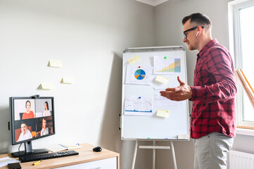 Fototapeta na wymiar Confident young man conducts webinar, business training online. Online coach stands near flip chart in front of PC display and explains something to online audience