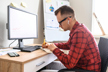 A concentrated guy is making notes into his notebook while sitting at the office desk in front of the computer, he is wearing glasses and headphones