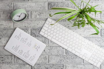 Top view on office desk with keyboard, paperclips, vase with plant and clock. Flat lay on gray brick background